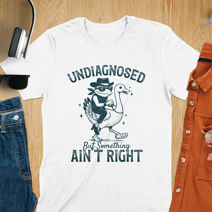 Funny cowboy frog riding goose t-shirt in white featuring cartoon frog in cowboy hat and boots riding a goose with text 'UNDIAGNOSED - But Something AIN'T RIGHT