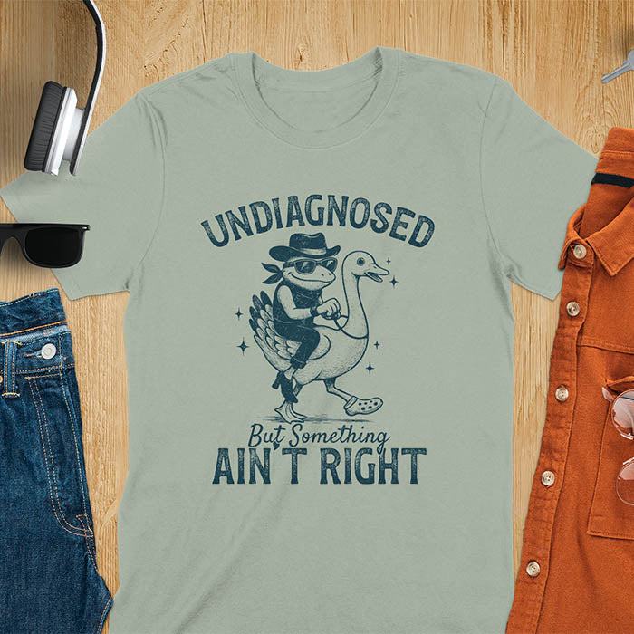 Funny cowboy frog riding goose t-shirt in sage featuring cartoon frog in cowboy hat and boots riding a goose with text 'UNDIAGNOSED - But Something AIN'T RIGHT