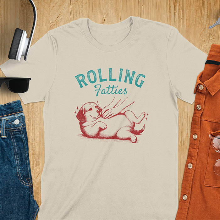 Sand colored t-shirt featuring 'Rolling Fatties' text in teal lettering above a chubby beagle dog lying on its back in red/burgundy ink sketch style