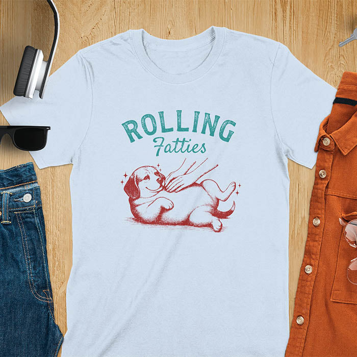 Light blue t-shirt featuring 'Rolling Fatties' text in teal lettering above a chubby beagle dog lying on its back in red/burgundy ink sketch style