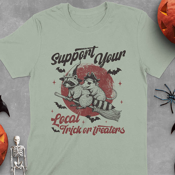 Sage green t-shirt Halloween witch raccoon design featuring opossum pirate on broom with Support Your Local Trick or Treaters text