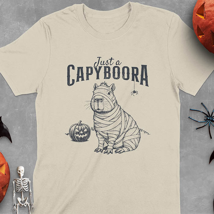 Sand colored t-shirt with a Halloween mummy capyboora t-shirt featuring cute capybara wrapped in white bandages with spooky Halloween design