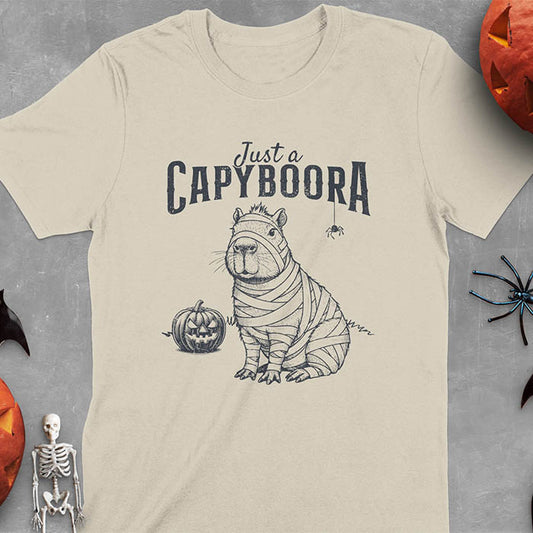 Sand colored t-shirt with a Halloween mummy capyboora t-shirt featuring cute capybara wrapped in white bandages with spooky Halloween design
