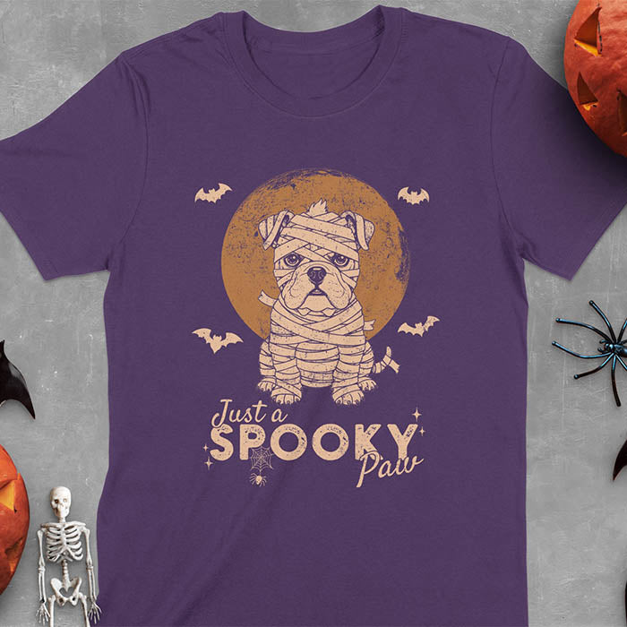 Purple t-shirt featuring adorable mummy bulldog puppy with white bandages and big eyes, Halloween design with 'Just a SPOOKY PAW' text below
