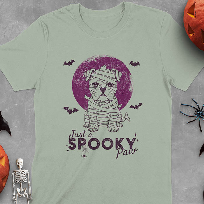 Sage green t-shirt featuring adorable mummy bulldog puppy with white bandages and big eyes, Halloween design with 'Just a SPOOKY PAW' text below