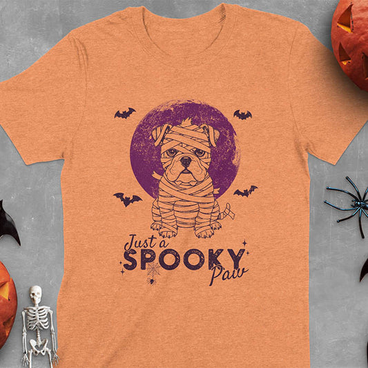 Heather Orange t-shirt featuring adorable mummy bulldog puppy with white bandages and big eyes, Halloween design with 'Just a SPOOKY PAW' text below