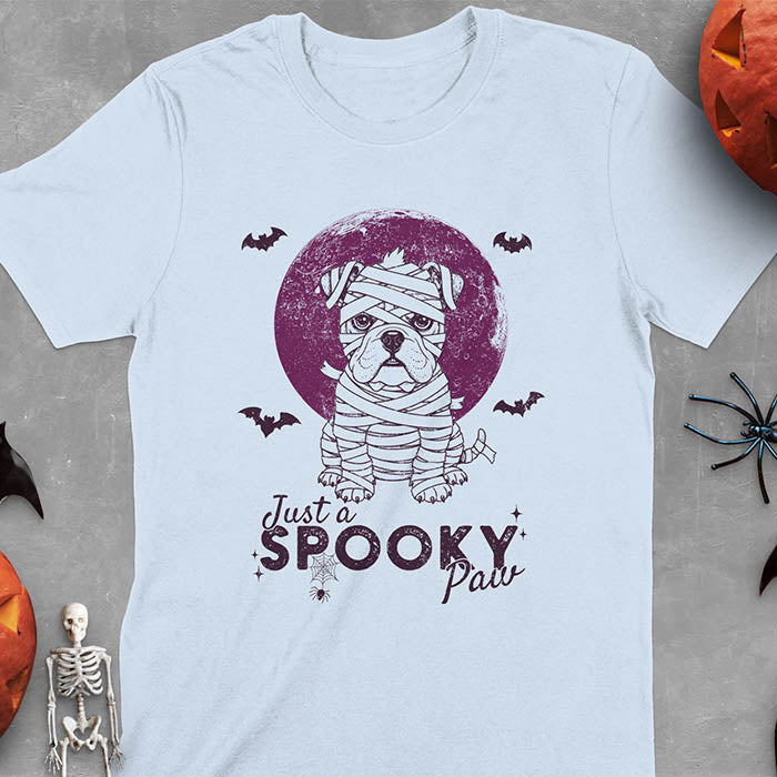 Light blue t-shirt featuring adorable mummy bulldog puppy with white bandages and big eyes, Halloween design with 'Just a SPOOKY PAW' text below