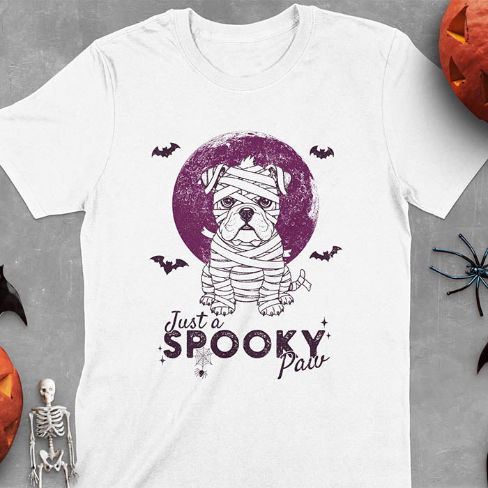 White t-shirt featuring adorable mummy bulldog puppy with white bandages and big eyes, Halloween design with 'Just a SPOOKY PAW' text below