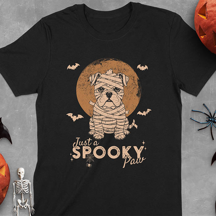 Black t-shirt featuring adorable mummy bulldog puppy with white bandages and big eyes, Halloween design with 'Just a SPOOKY PAW' text below