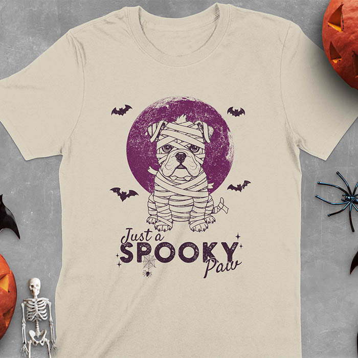 Sand colored t-shirt featuring adorable mummy bulldog puppy with white bandages and big eyes, Halloween design with 'Just a SPOOKY PAW' text below