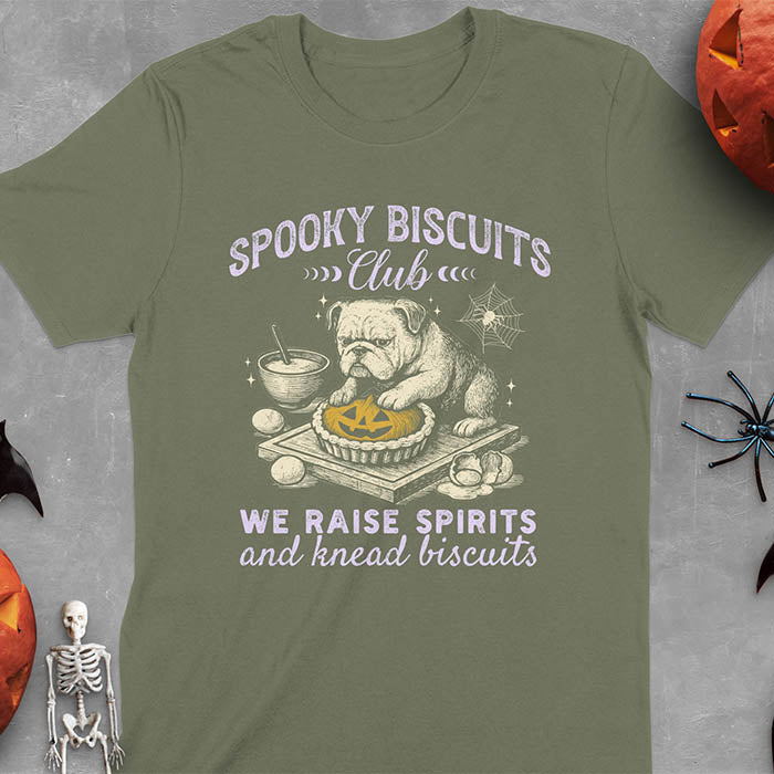 Military Green t-shirt featuring 'Spooky Biscuits Club' design with illustrated bulldog sitting at wooden table with Halloween pumpkin-shaped biscuit, mixing bowl, eggs, and spider web decoration, text reads 'We raise spirits and knead biscuits'