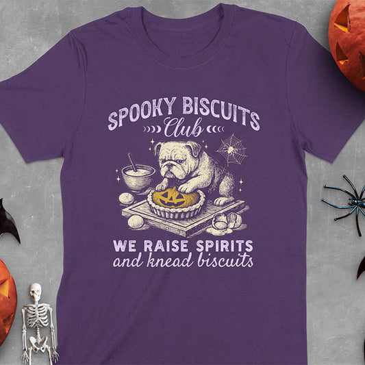 Purple t-shirt featuring 'Spooky Biscuits Club' design with illustrated bulldog sitting at wooden table with Halloween pumpkin-shaped biscuit, mixing bowl, eggs, and spider web decoration, text reads 'We raise spirits and knead biscuits'