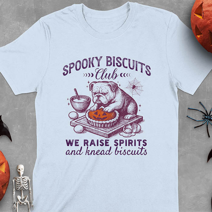 Light blue t-shirt featuring 'Spooky Biscuits Club' design with illustrated bulldog sitting at wooden table with Halloween pumpkin-shaped biscuit, mixing bowl, eggs, and spider web decoration, text reads 'We raise spirits and knead biscuits'