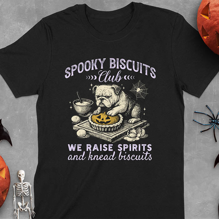 Black t-shirt featuring 'Spooky Biscuits Club' design with illustrated bulldog sitting at wooden table with Halloween pumpkin-shaped biscuit, mixing bowl, eggs, and spider web decoration, text reads 'We raise spirits and knead biscuits'