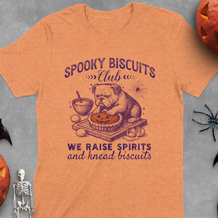Heather Orange t-shirt featuring 'Spooky Biscuits Club' design with illustrated bulldog sitting at wooden table with Halloween pumpkin-shaped biscuit, mixing bowl, eggs, and spider web decoration, text reads 'We raise spirits and knead biscuits'