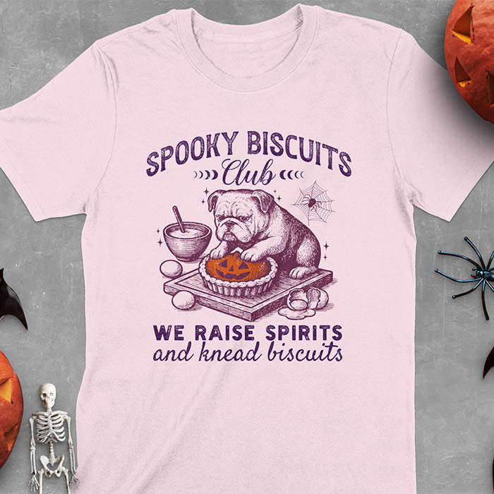 Light pink t-shirt featuring 'Spooky Biscuits Club' design with illustrated bulldog sitting at wooden table with Halloween pumpkin-shaped biscuit, mixing bowl, eggs, and spider web decoration, text reads 'We raise spirits and knead biscuits'