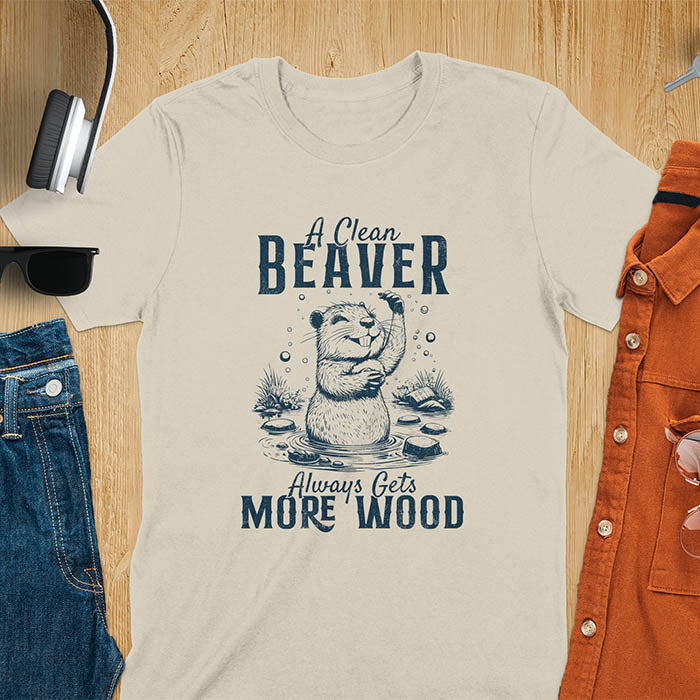 Sand colored t-shirt featuring illustrated beaver bathing in water with text 'A Clean Beaver Always Gets More Wood' - adult humor meme design with cartoon beaver surrounded by water bubbles and tree stumps