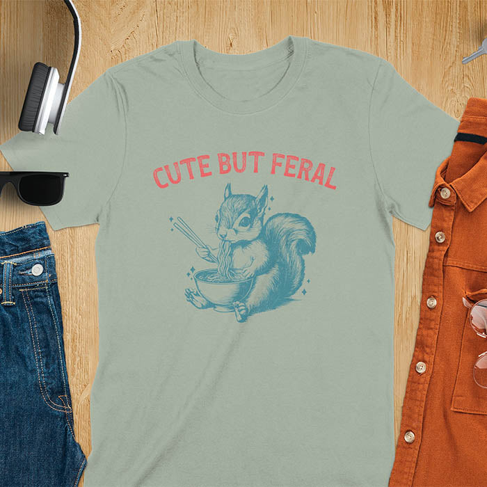 Sage Green t-shirt featuring illustrated squirrel holding chopsticks and eating from ramen bowl with red text 'CUTE BUT FERAL' above - playful animal meme design