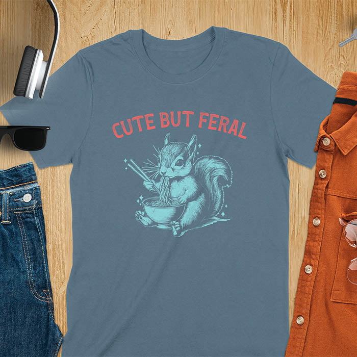 Indigo Blue t-shirt featuring illustrated squirrel holding chopsticks and eating from ramen bowl with red text 'CUTE BUT FERAL' above - playful animal meme design