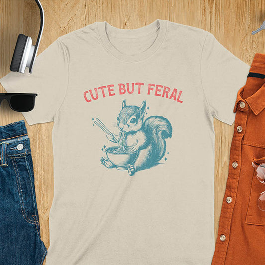 Sand color t-shirt featuring illustrated squirrel holding chopsticks and eating from ramen bowl with red text 'CUTE BUT FERAL' above - playful animal meme design