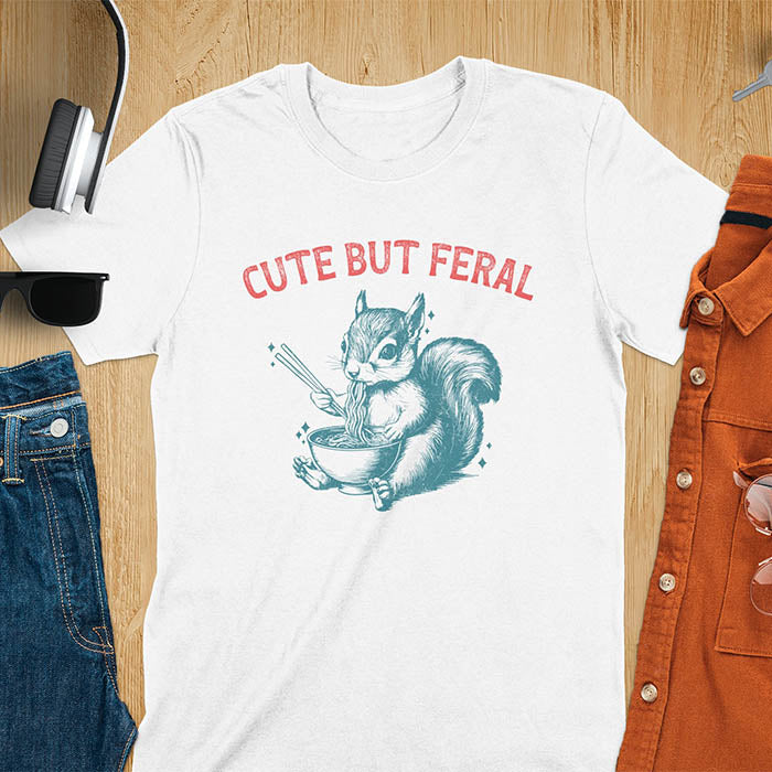 White t-shirt featuring illustrated squirrel holding chopsticks and eating from ramen bowl with red text 'CUTE BUT FERAL' above - playful animal meme design