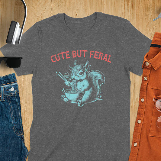 Dark heather t-shirt featuring illustrated squirrel holding chopsticks and eating from ramen bowl with red text 'CUTE BUT FERAL' above - playful animal meme design