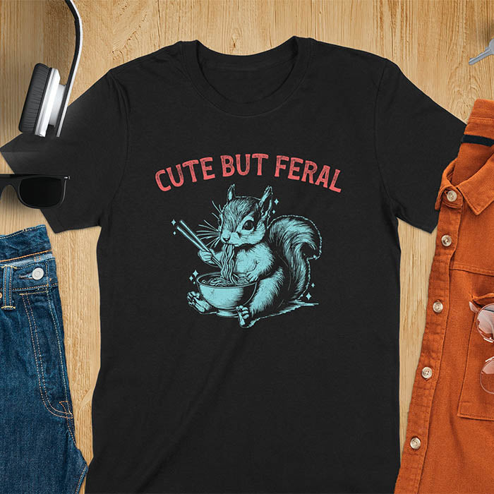 Black t-shirt featuring illustrated squirrel holding chopsticks and eating from ramen bowl with red text 'CUTE BUT FERAL' above - playful animal meme design
