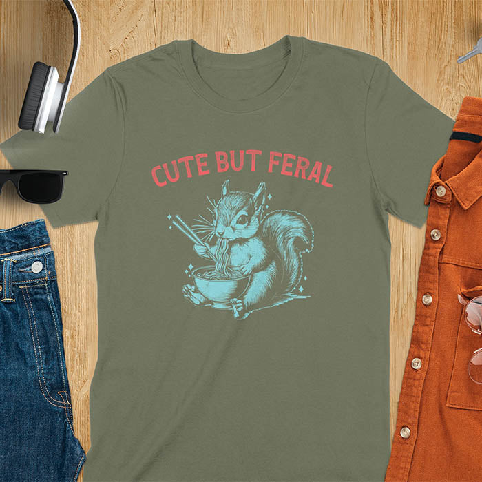 Military Green t-shirt featuring illustrated squirrel holding chopsticks and eating from ramen bowl with red text 'CUTE BUT FERAL' above - playful animal meme design