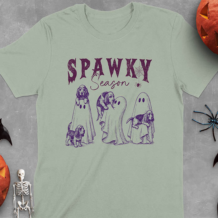 Sage green Halloween T-shirt featuring 'SPAWKY Season' text with cute ghosts carrying Basset Hound puppies, designed in purple ink with playful Halloween theme