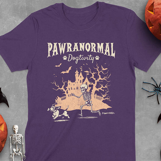 Purple Halloween T-shirt featuring 'PAWRANORMAL DOGTIVITY' text with humorous scene of Basset Hound running away with a skeleton bone while being chased by an angry one-armed skeleton with a haunted house background
