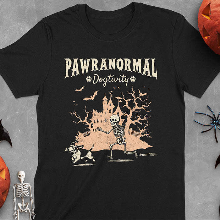 Black Halloween T-shirt featuring 'PAWRANORMAL DOGTIVITY' text with humorous scene of Basset Hound running away with skeleton bones while being chased by an angry one-armed skeleton with a haunted house background