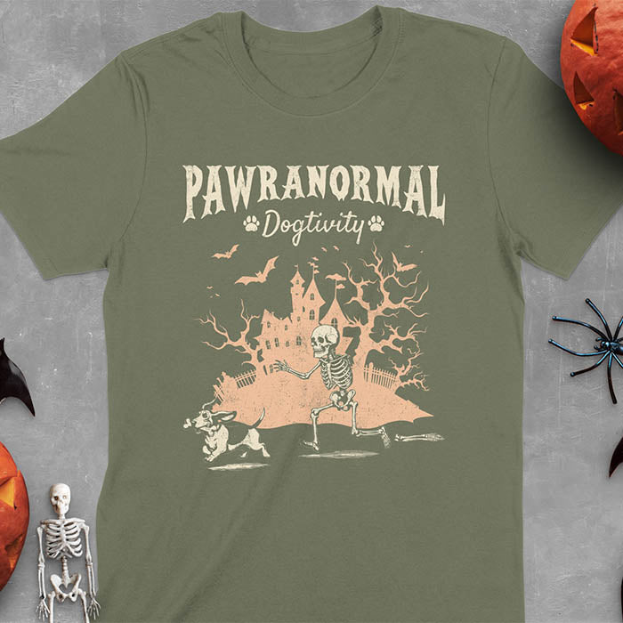 Military Green Halloween T-shirt featuring 'PAWRANORMAL DOGTIVITY' text with humorous scene of Basset Hound running away with skeleton bones while being chased by an angry one-armed skeleton with a haunted house background