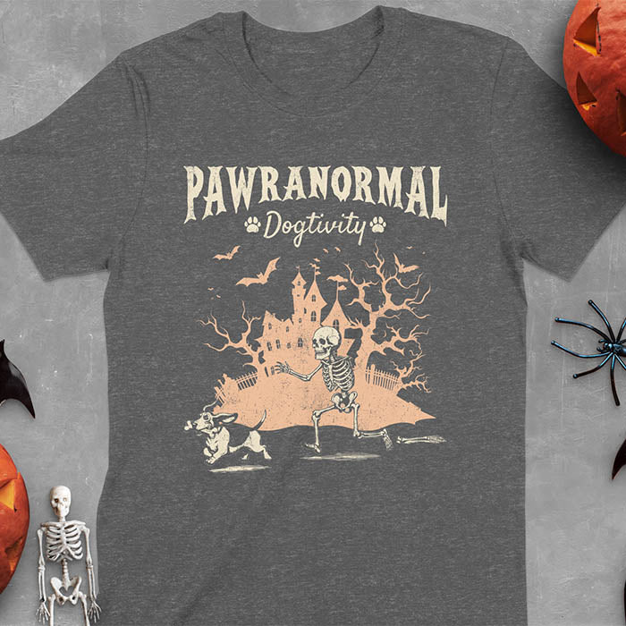 Dark heather Halloween T-shirt featuring 'PAWRANORMAL DOGTIVITY' text with humorous scene of Basset Hound running away with skeleton bones while being chased by an angry one-armed skeleton with a haunted house background