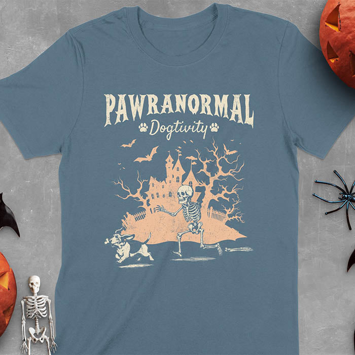 Indigo Blue Halloween T-shirt featuring 'PAWRANORMAL DOGTIVITY' text with humorous scene of Basset Hound running away with skeleton bones while being chased by an angry one-armed skeleton with a haunted house background