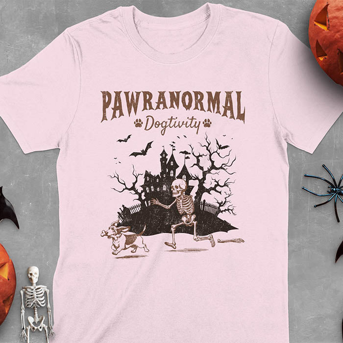 Light pink Halloween T-shirt featuring 'PAWRANORMAL DOGTIVITY' text with humorous scene of Basset Hound running away with skeleton bones while being chased by an angry one-armed skeleton, designed in brown ink with a haunted house background