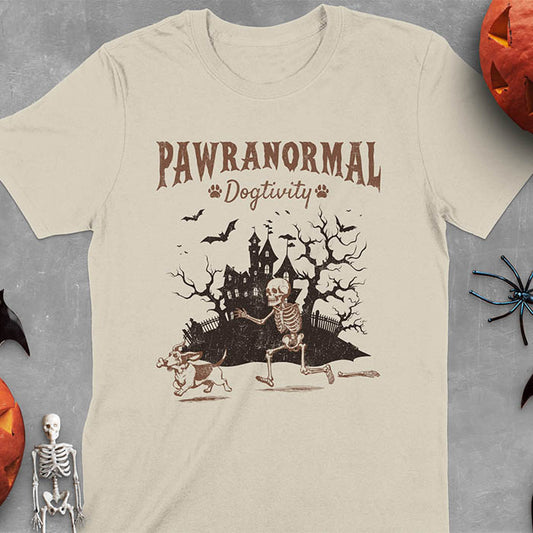 Sand colored Halloween T-shirt featuring 'PAWRANORMAL DOGTIVITY' text with humorous scene of Basset Hound running away with skeleton bones while being chased by an angry one-armed skeleton, designed in brown ink with a haunted house background