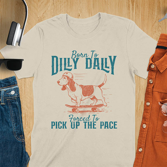 Basset hound wearing helmet on skateboard with Born To Dilly Dally Forced To Pick Up The Pace lettering on sand color tee