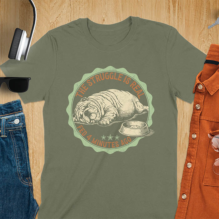 Military Green t-shirt with vintage badge graphic of a chubby English bulldog lying next to an empty food bowl, text reads "The Struggle Is Real Fed 4 Minutes Ago"