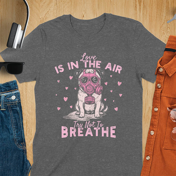 Anti-Valentine's Day pug wearing pink gas mask graphic tee with Love is in the Air Try Not to Breathe text and hearts on dark heather shirt