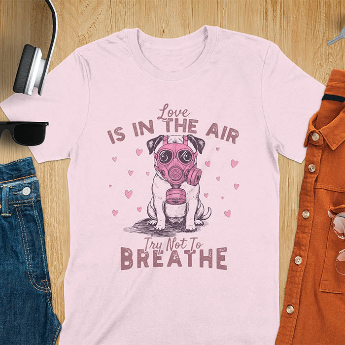 Anti-Valentine's Day pug wearing pink gas mask graphic tee with Love is in the Air Try Not to Breathe text and hearts on light pink shirt