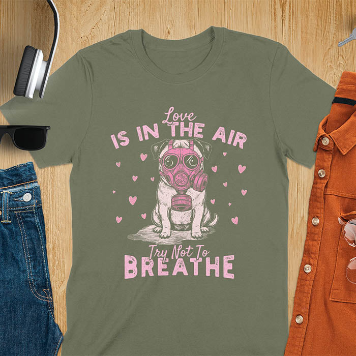 Anti-Valentine's Day pug wearing pink gas mask graphic tee with Love is in the Air Try Not to Breathe text and hearts on military green shirt