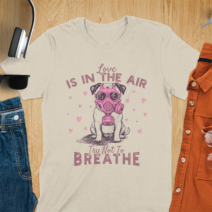 Anti-Valentine's Day pug wearing pink gas mask graphic tee with Love is in the Air Try Not to Breathe text and hearts on sand color shirt