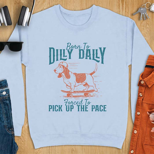 Light Blue sweatshirt with a graphic of a basset hound in a skater helmet riding a skateboard with long ears flying back, printed with the text Born To Dilly Dally, Forced To Pick Up The Pace