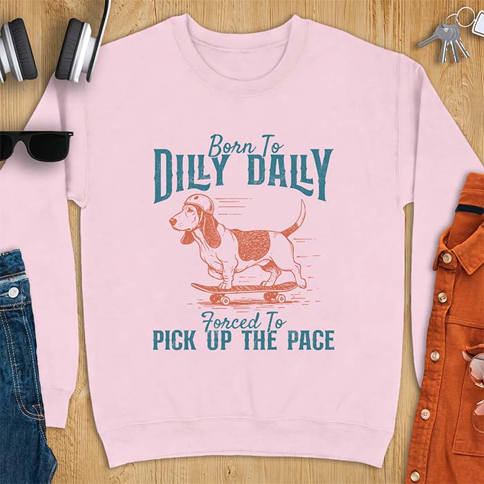 Light Pink sweatshirt with a graphic of a basset hound in a skater helmet riding a skateboard with long ears flying back, printed with the text Born To Dilly Dally, Forced To Pick Up The Pace