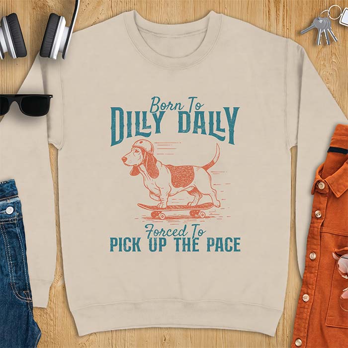 Sand sweatshirt with a graphic of a basset hound in a skater helmet riding a skateboard with long ears flying back, printed with the text Born To Dilly Dally, Forced To Pick Up The Pace