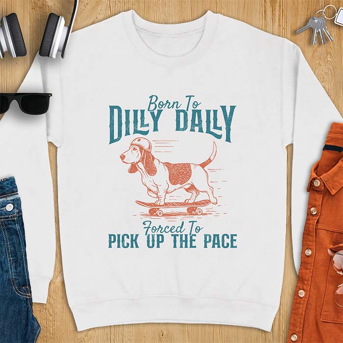 White sweatshirt with a graphic of a basset hound in a skater helmet riding a skateboard with long ears flying back, printed with the text Born To Dilly Dally, Forced To Pick Up The Pace