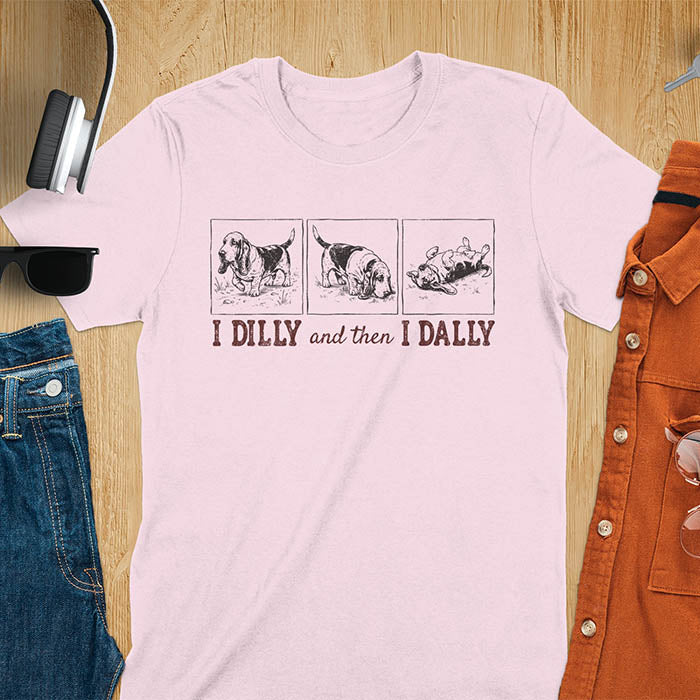 Basset hound comic strip tee — three panels: walking, sniffing, flopped over — with text "I DILLY and then I DALLY" in bold retro typography, light pink