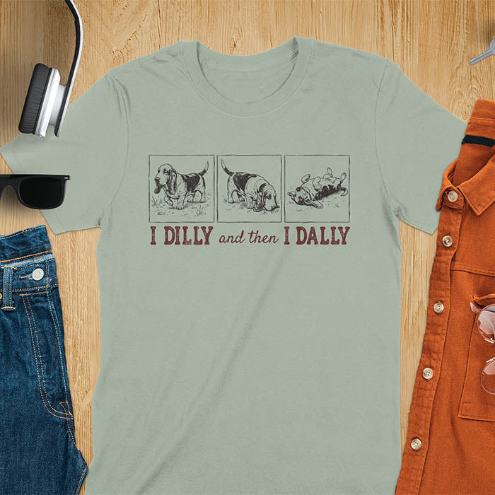 Basset hound comic strip tee — three panels: walking, sniffing, flopped over — with text "I DILLY and then I DALLY" in bold retro typography, sage green