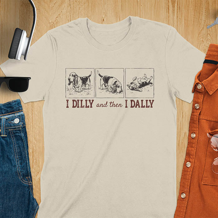 Basset hound comic strip tee — three panels: walking, sniffing, flopped over — with text "I DILLY and then I DALLY" in bold retro typography, sand color