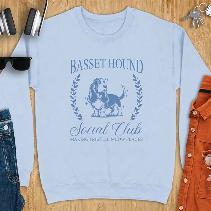 Light Blue sweatshirt with a vintage crest graphic of a basset hound standing inside a laurel wreath and the text Basset Hound, Social Club and Making Friends In Low Places
